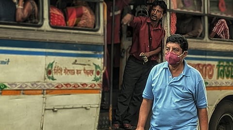The image showing a crowded bus with Hindi text "Prem Seva Bus Transport" and colorful patterns, with passengers boarding. A man in a blue shirt and light pants walking in the foreground.