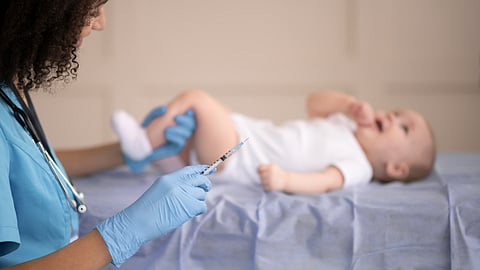 A boy baby on an examination table and a nurse approaching with a syringe.
