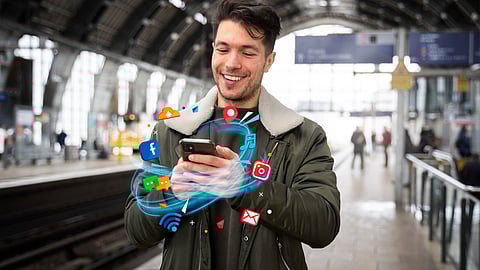 A man on a train platform uses a smartphone with social media and app icons overlaid around his hands, set against a blurred station background.