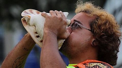 Image of a man having curly hairs and wearing glasses and have a tattoo blowing a conch shell.
