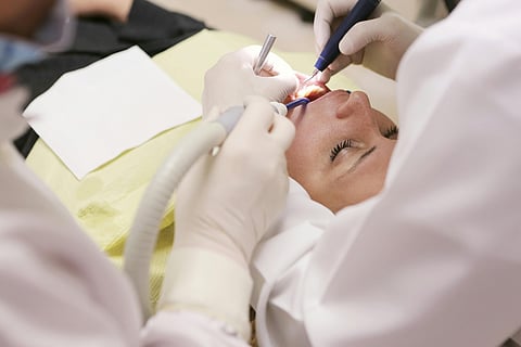 A woman sits in a dental chair while a dentist cleans her teeth with dental tools.