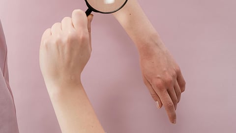 Image of a woman checking hands of another woman using a microscope in front of pink background.