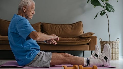 An elderly man is doing some exercises at his home sitting on a mat on the floor and a pair of dumbbells kept nearby.