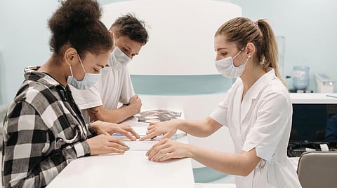 Three masked individuals in a dental office, engaged in a conversation near dental equipment.