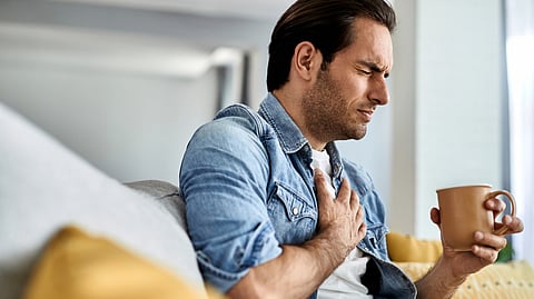Young man feeling sick and holding his chest in pain while drinking tea in the living room.