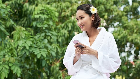 An Asian looking woman wearing a white robe and a flower on her ears sitting outside.