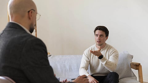 A young man sitting on a couch is speaking to another man wearing a black suit and sitting in a chair opposite to him.  