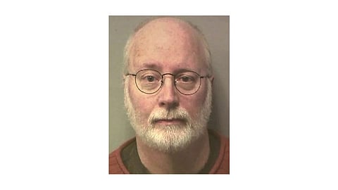 An older man with glasses and a beard is depicted in a mug shot, facing the camera with a neutral expression.
