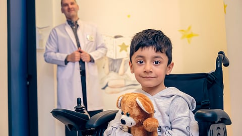 A boy in a wheelchair smiles while holding a teddy bear close to his chest.