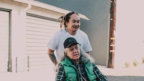 An image of a young person standing behind a seated old person.