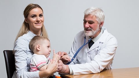 A male doctor is examining a kid sitting in his mother's lap with a stethoscope. 