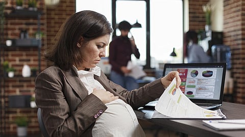 A pregnant woman wearing a brown blazer is looking at some papers at workplace.