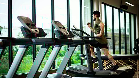A man exercising on a treadmill in a gym, surrounded by fitness equipment and bright overhead lights.