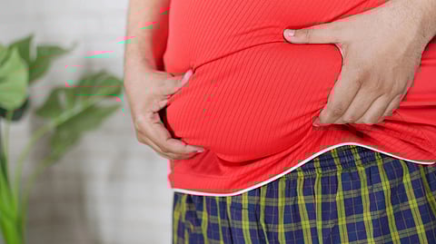 Close-up image of a fat person wearing a red t-shirt and checkered joggers holding his belly.