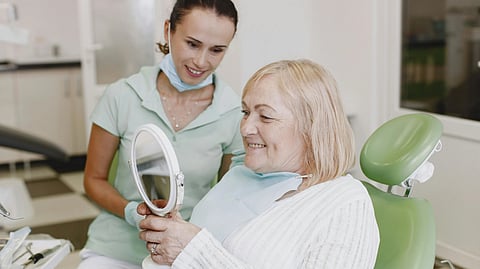 A woman sits in a dental chair while another woman examines her reflection in a mirror.