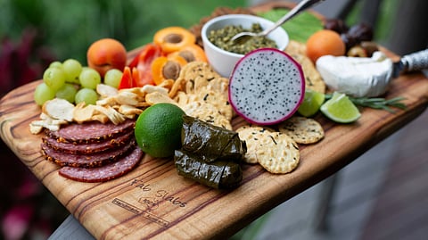 A wooden cutting board topped with assorted foods, including vegetables, fruits, and meats, arranged for preparation.