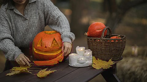 A woman carves a pumpkin on a picnic table, surrounded by autumn leaves and tools for pumpkin decoration.