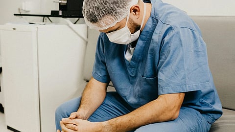 An image of man in scrubs sits on a bench, appearing relaxed and contemplative in a medical environment.