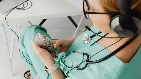 A woman in scrubs sits at a desk and holding a bottle of tablets.