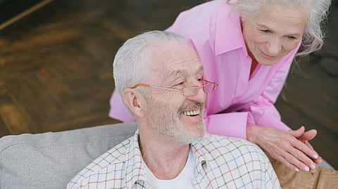 Image of a woman wearing a pink shirt standing behind an old man wearing a white checkered shirt and sitting on a sofa.