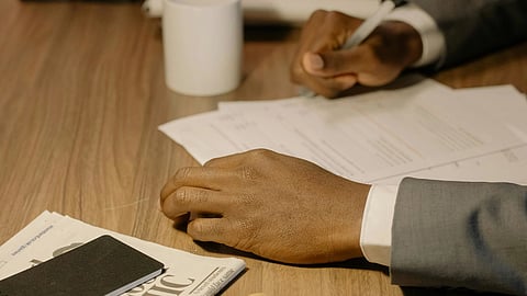 A man in a suit sits at a desk, surrounded by papers and holding a pen, focused on his work.