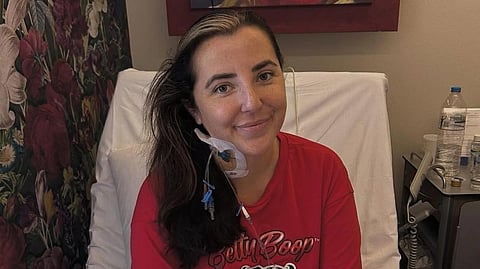 An image of Lisa Walsh post-surgery sitting on a bed in a red tshirt.