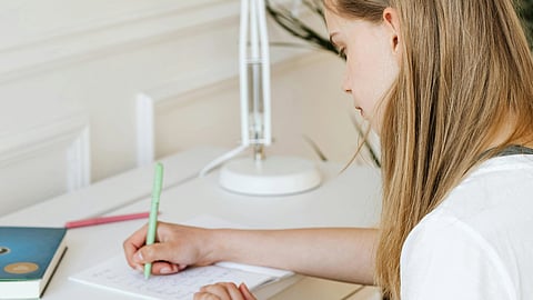  A girl sits at a desk, writing in a notebook with a pen, focused on her work.