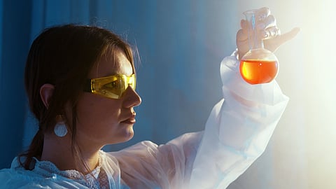 Image of a scientist wearing yellow safety glasses examines an orange liquid in a flask.