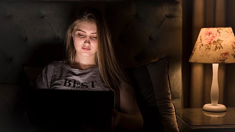 Young woman in a dimly lit room sits on a bed, focused on a laptop. Soft lamp light creates a cozy, calm ambiance. Keywords: night, alone, focused.