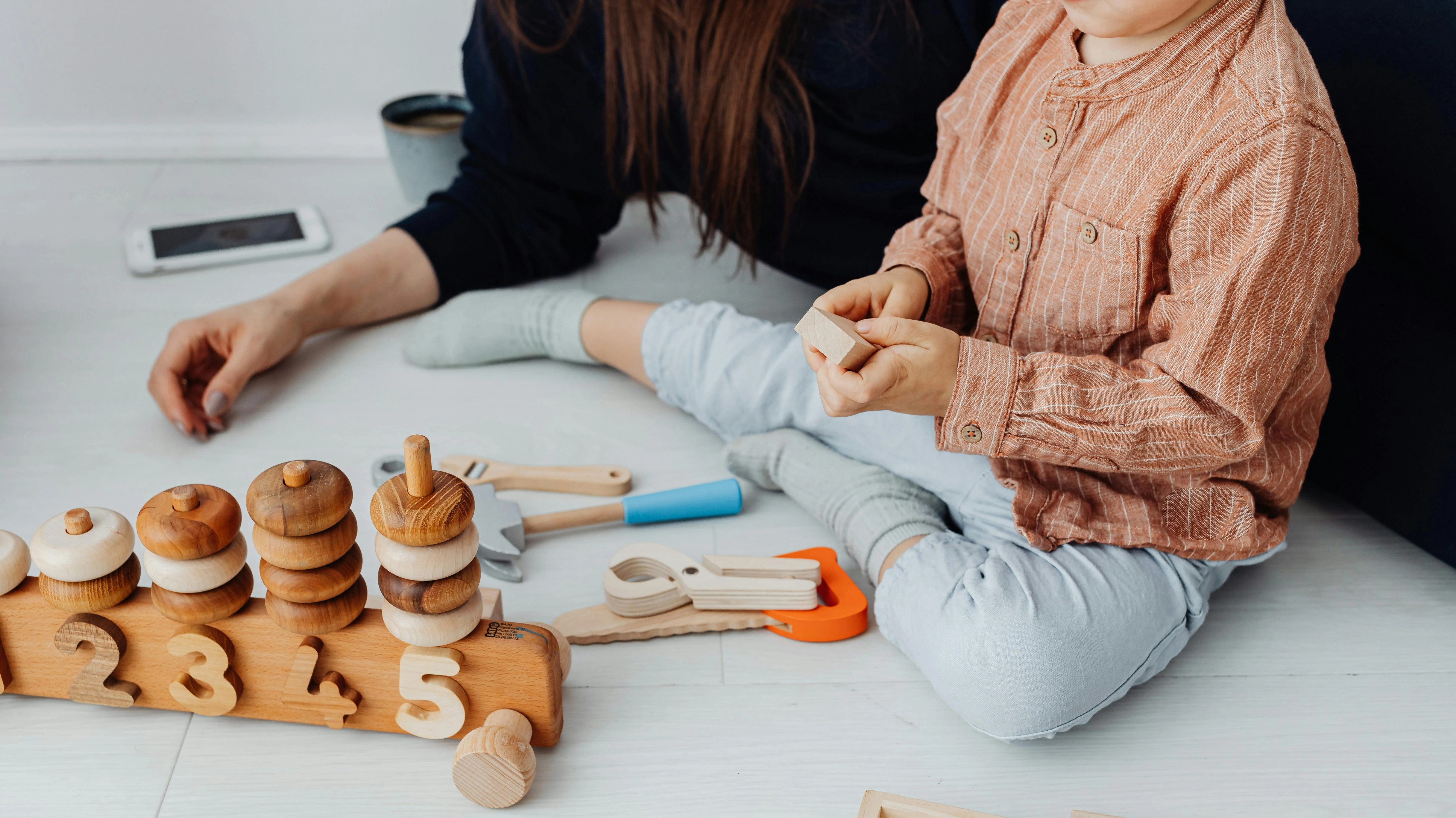 A woman and a child sit on the floor, playing with colorful wooden toys scattered around them.