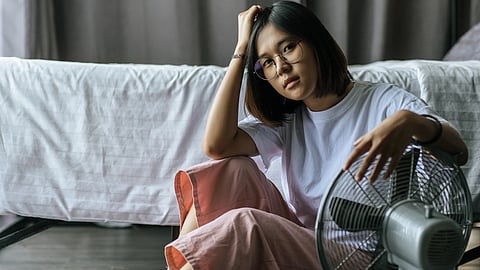 Image of a woman sitting on the floor beside a fan, looking relaxed in a casual setting.