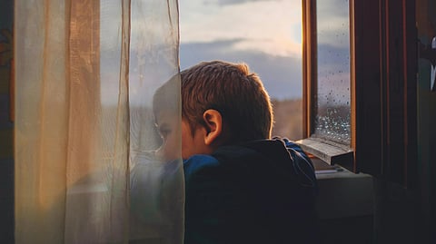 A child peers thoughtfully out an open window, with soft sunlight and sheer curtains creating a warm, contemplative ambiance.