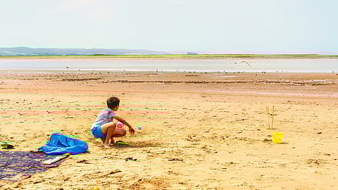 Image of a child in blue shorts sits on a sandy beach near a picnic blanket and toys, gazing towards the sea. 