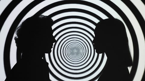 Two people stand together in front of a large black and white spiral design, creating a striking visual contrast.