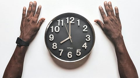 A person seated at a table, hands resting on a clock, indicating a moment of contemplation or time management.