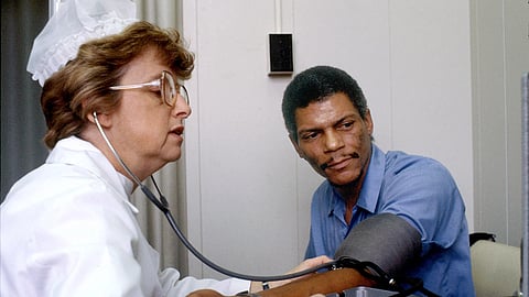 A white nurse in a white uniform, headgear, and spectacles checks a patient's blood pressure using a grey blood pressure cuff and a stethoscope.