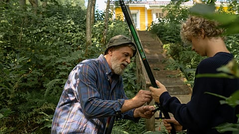 A man and a boy in the woods examine a fishing rod, surrounded by trees and natural scenery.