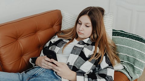 Image of a young girl in a plaid shirt and jeans reclines on a brown couch, focused on her smartphone, conveying a relaxed and absorbed mood.