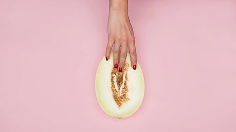 A hand with red nails rests on a halved melon on a pink background.
