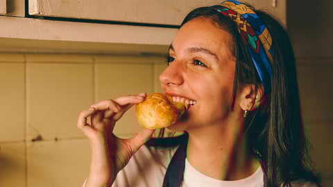 Image of a woman wearing a blue headband, standing and eating bun in the kitchen.