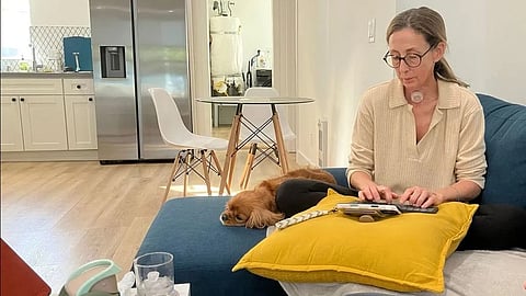 A woman wearing glasses is seated on a blue couch, using a tablet. A brown dog rests beside her on a yellow pillow. 