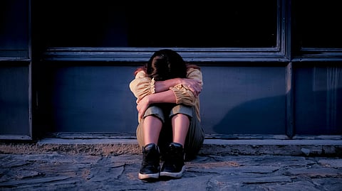 A person sits on a stone sidewalk against a dark wall, head resting on folded arms and knees.