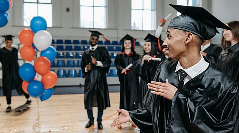 Graduates in caps and gowns celebrate indoors, smiling and clapping, near balloons and confetti on the floor. 