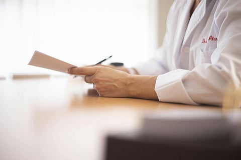 A doctor's left hand holding a pen and paper on the wooden table