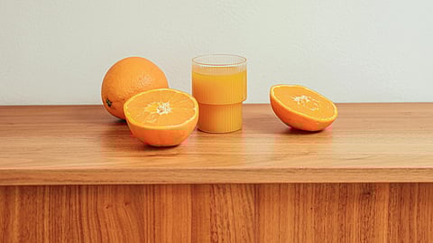 Glass of orange juice and two whole oranges sit on a wooden tray. 