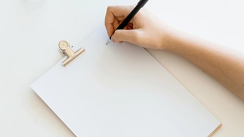 Woman writing on blank paper with a pen on a white table, focused on her task.