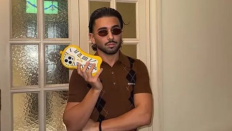 A man wearing a brown shirt and pants holds a banana, smiling at the camera.