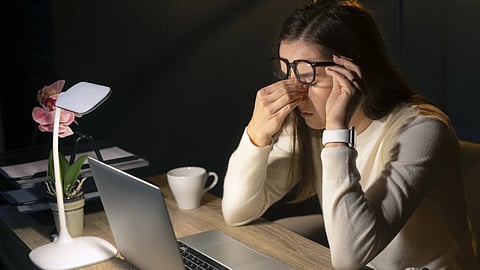 An Asian woman rubbing her eyes sitting in front of a laptop with a table lamp.