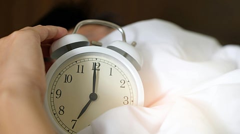 Image of a white alarm clock showing 7:00 and is on a pink sheet.