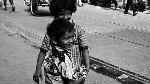 A black-and-white image of two children wearing shirts, smiling as they stand on a street.